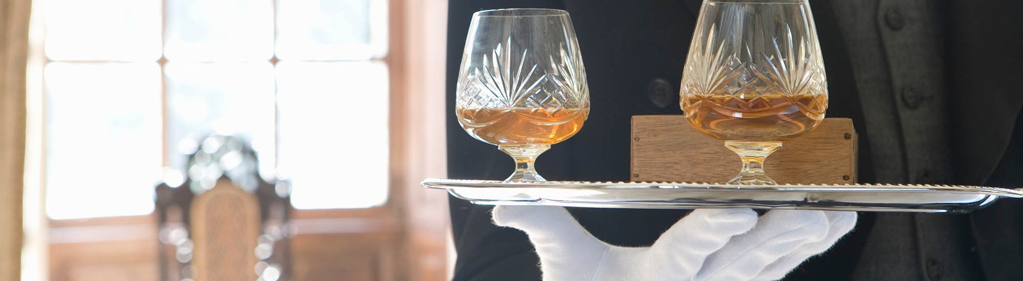 Close up of a tray containing two glasses of whiskey carried by a butler inside a luxurious house on a bright, sunny day