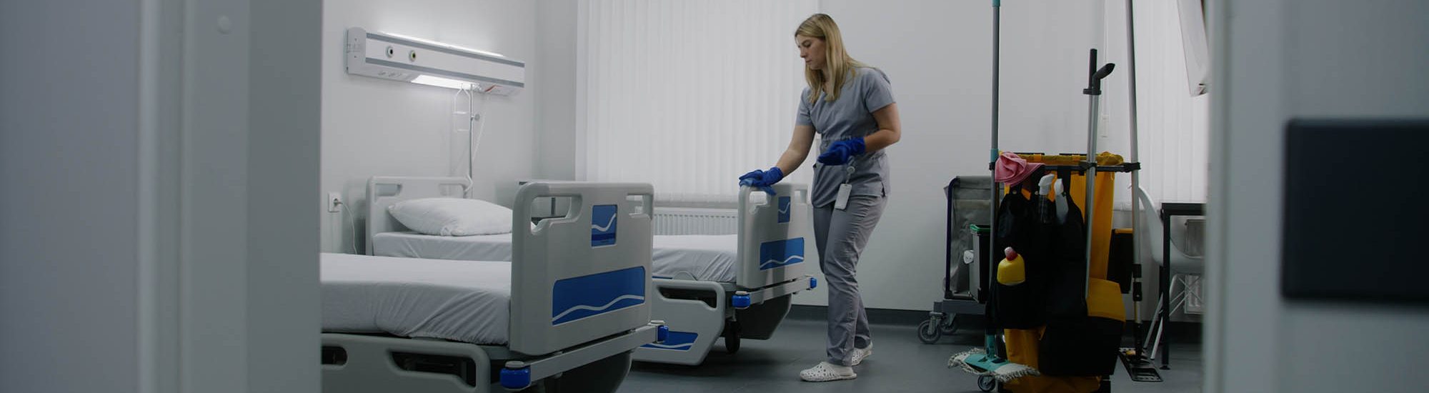Adult nurse mops floor between beds in hospital ward. Health worker brings cleaning trolley to hospital room. Female cleaner prepares ward for new patients. Medical staff at work in modern clinic.