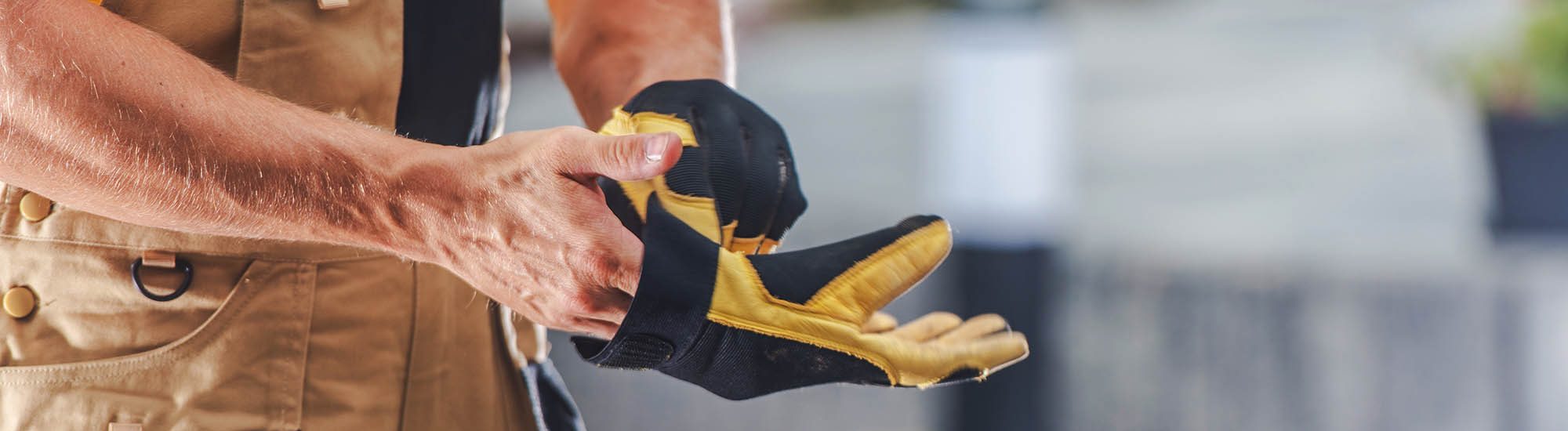 A construction worker puts on safety gloves, preparing for a task in a vibrant outdoor work area filled with greenery and tools. The worker is focused and diligent.
